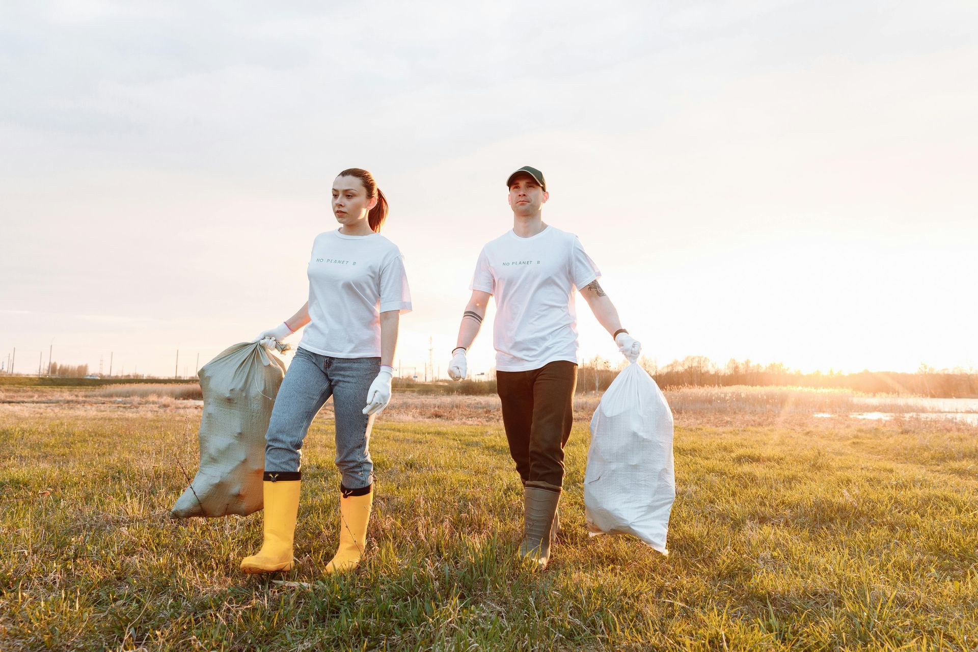 A man and a woman are walking through a grassy field holding garbage bags.