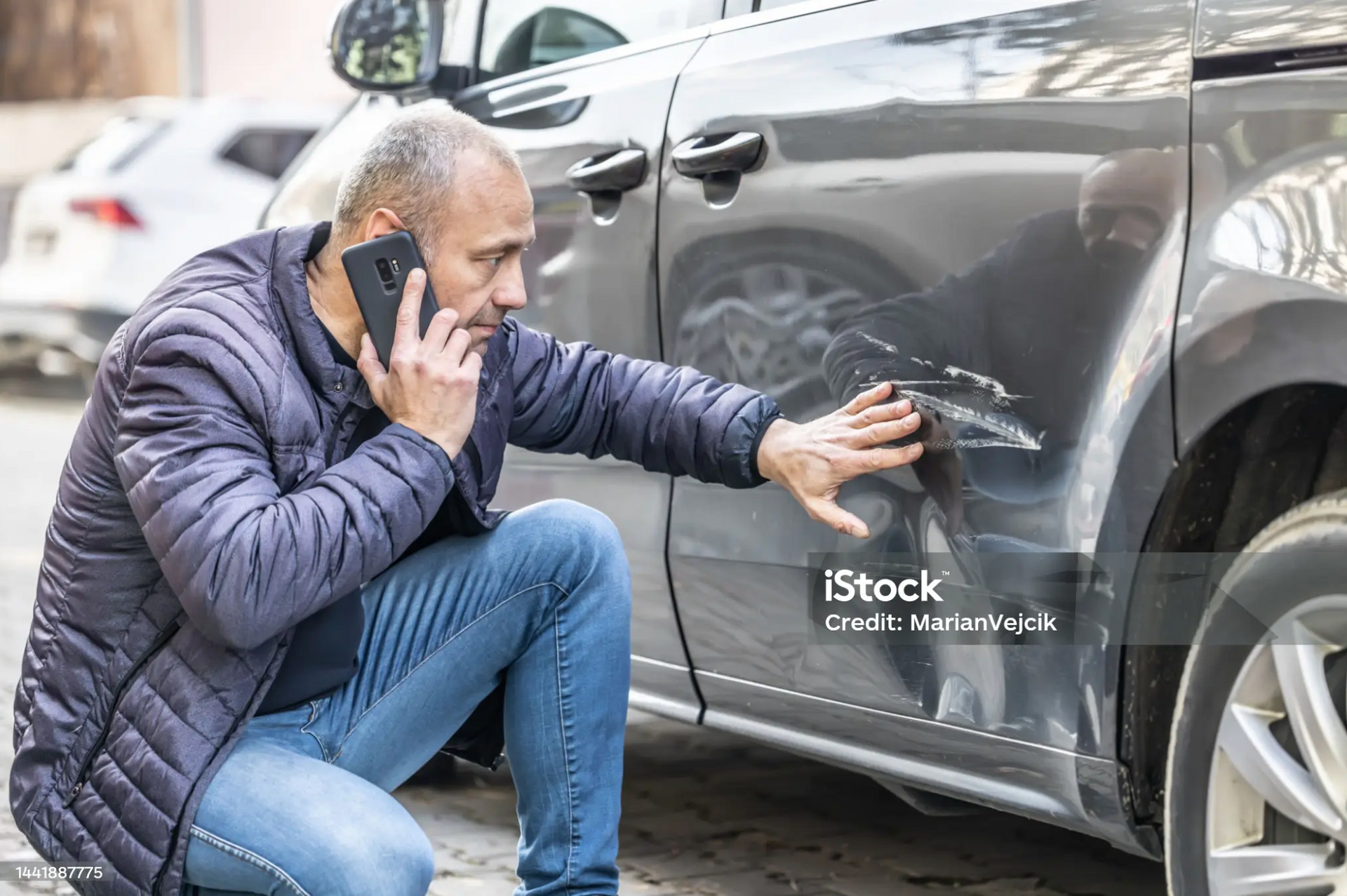 Man inspecting a scratch on the side of a car while speaking on the phone to his insurance company.