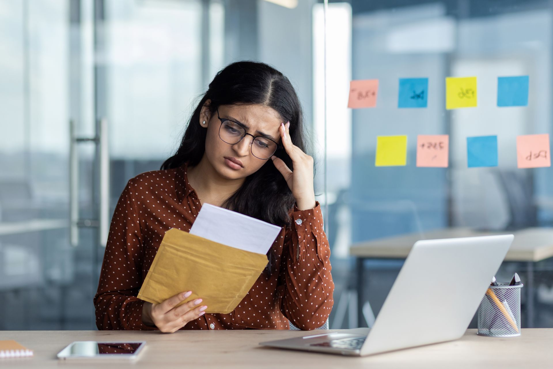 A woman sitting at a desk looking frustrated while holding a denial letter, with a laptop and sticky