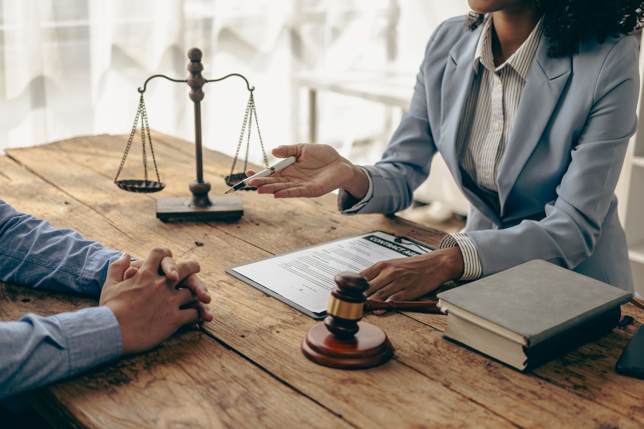 A lawyer in a suit discussing a legal document with a client across the table, with scales of justice and a gavel on the desk.