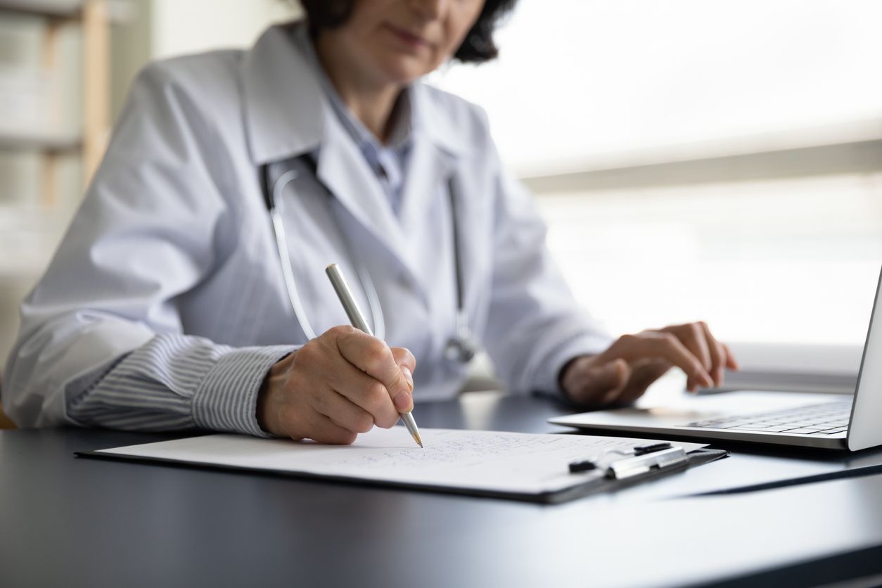 A doctor writes medical notes on a clipboard while reviewing information on a laptop, highlighting the importance of strong medical evidence to be used by a long-term disability lawyer.