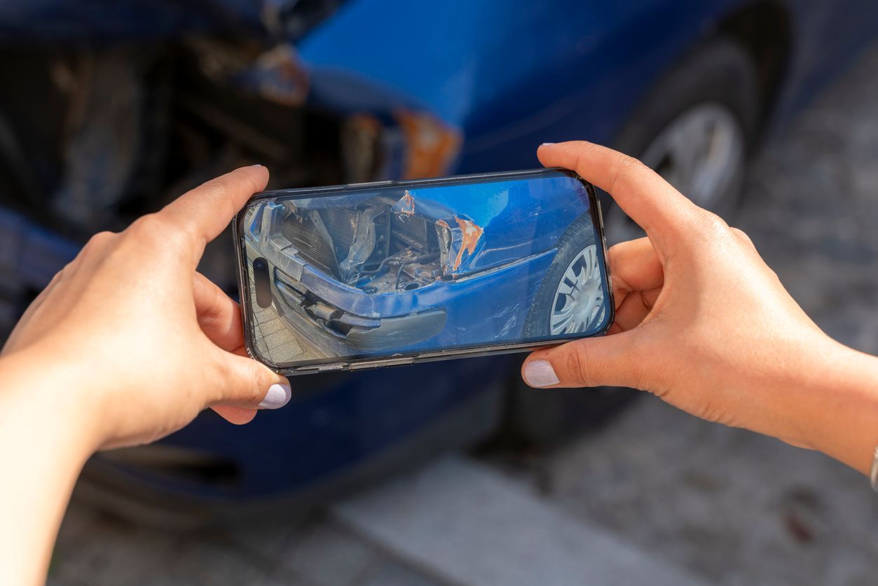 Hands holding a smartphone to photograph the damage on a blue car after an accident, highlighting the importance of documenting evidence.