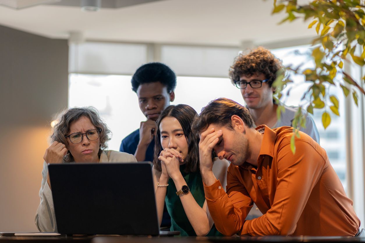 Group of concerned employees gathered around a laptop, discussing workplace layoffs in an office set