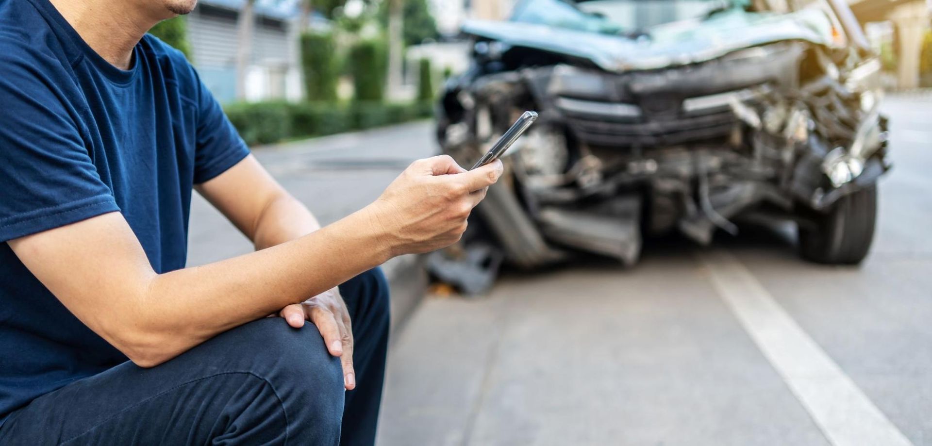 Man sitting on his phone with a wrecked car behind him.