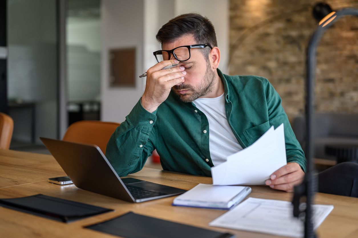 A man, sitting at his desk in front of his laptop and paperwork.
