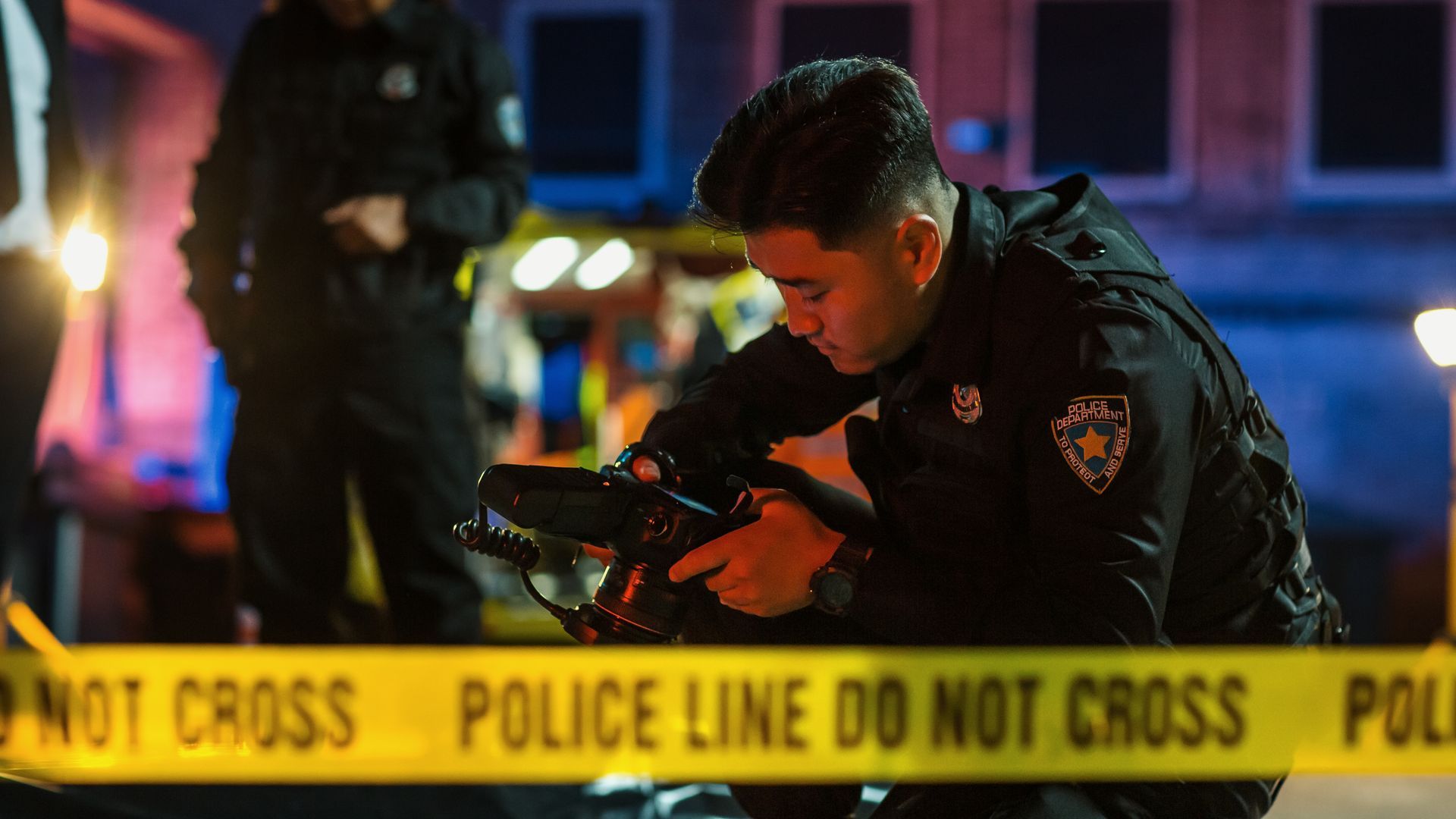 A police officer documenting evidence at a car accident scene, with a yellow Police Line Do Not Cross tape in the foreground.