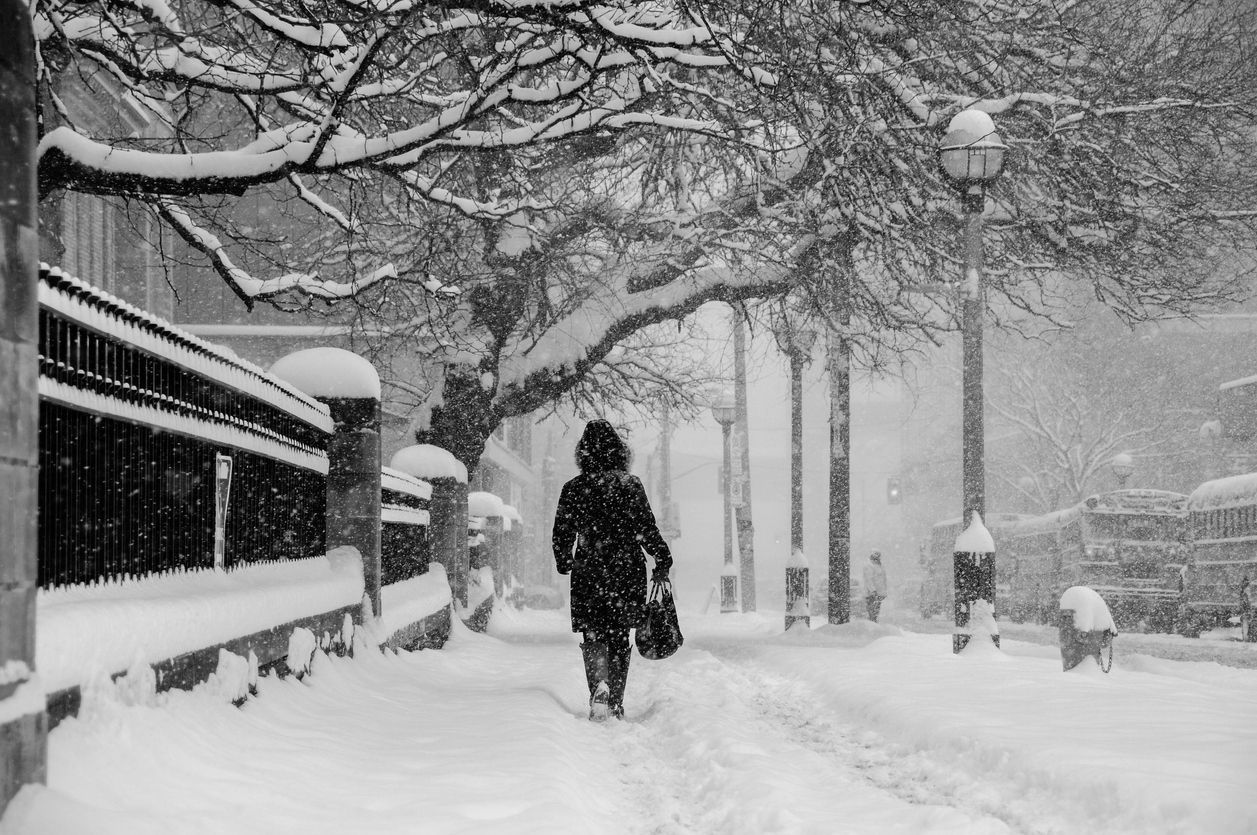 A person walking on a snow-covered sidewalk during a winter storm, highlighting slippery and hazardous conditions.