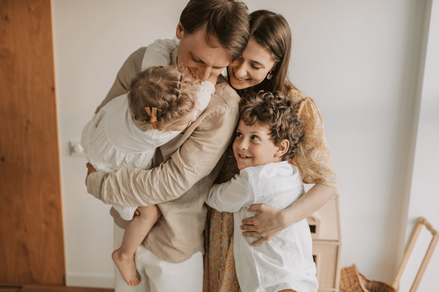 A family hugging in a bright room, with two adults and two children smiling closely together.