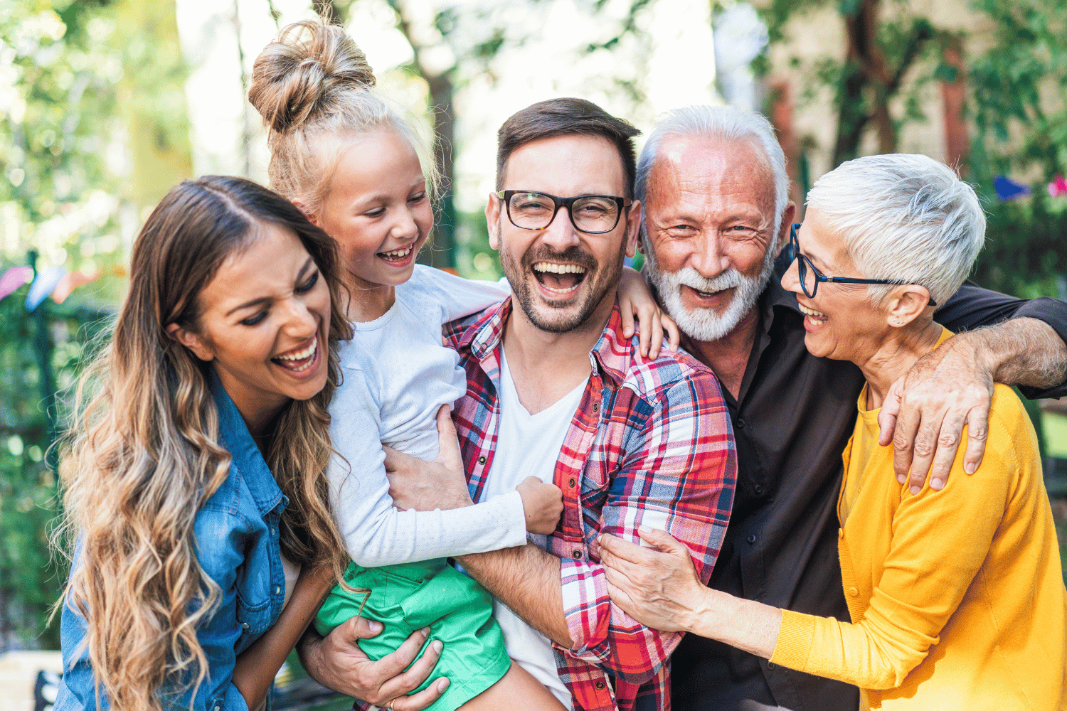 Five smiling people hugging outdoors, including a man in glasses and a woman in a yellow shirt