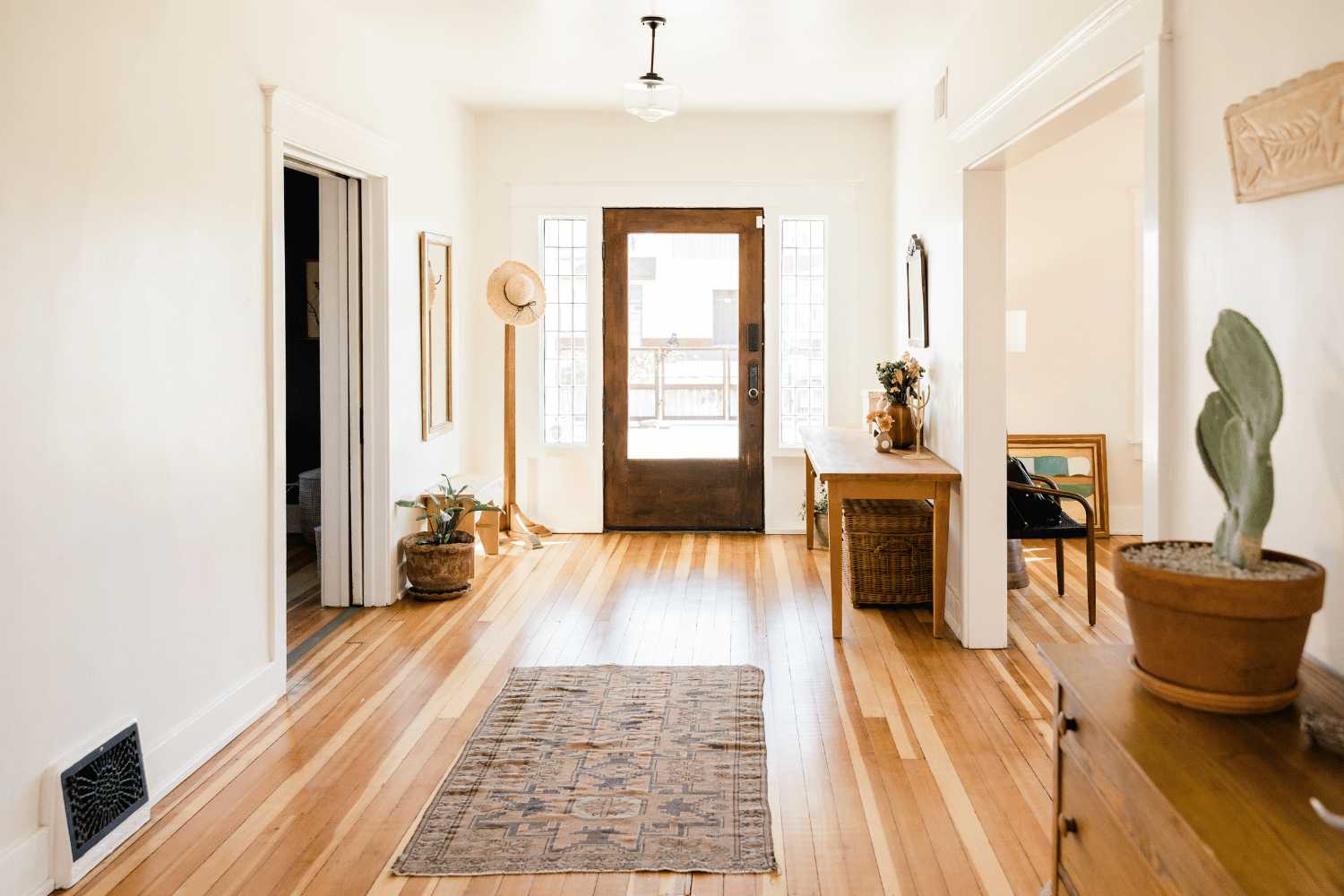 Bright entryway with hardwood floors, runner rug, wooden front door, and potted plants.