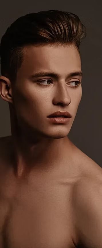 Ein Mann mit kurzem, gestyltem braunem Haar blickt aus dem Bild. Er hat freie Schultern und einen neutralen Gesichtsausdruck.