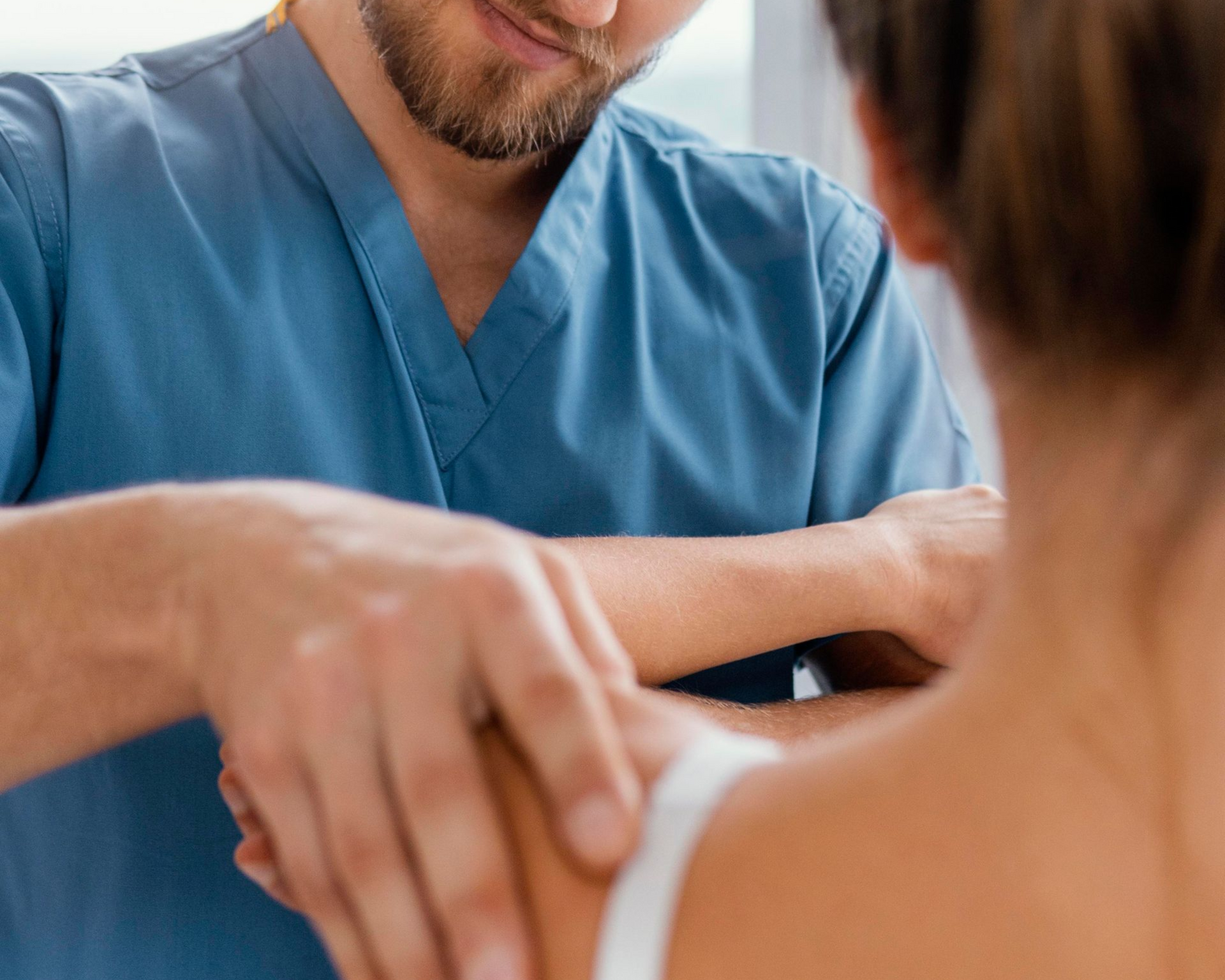 Healthcare worker in blue scrubs examining a patient’s shoulder with hands on the upper arm