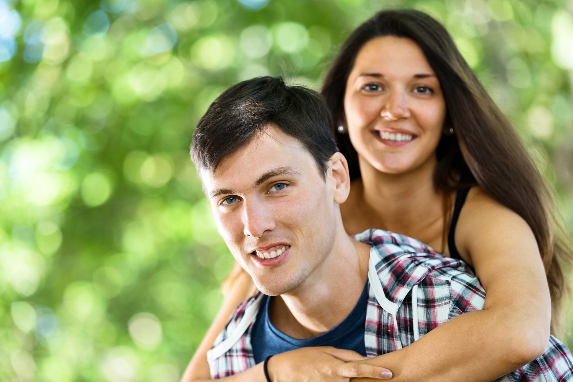 Smiling couple outdoors, woman hugging man from behind with green bokeh background