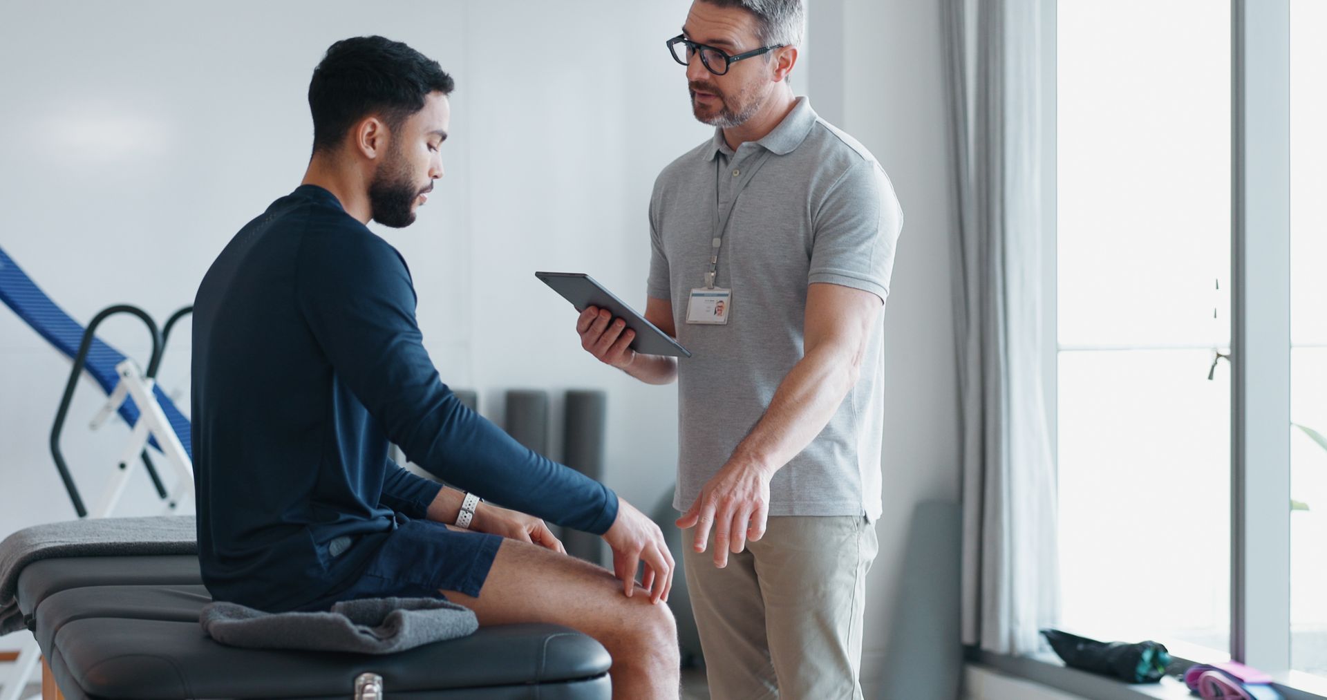 Therapist speaking with seated patient in a bright clinic, holding a tablet.