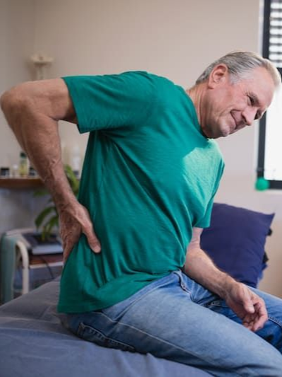Man in green shirt holding his lower back while sitting on a bed, appearing to have back pain
