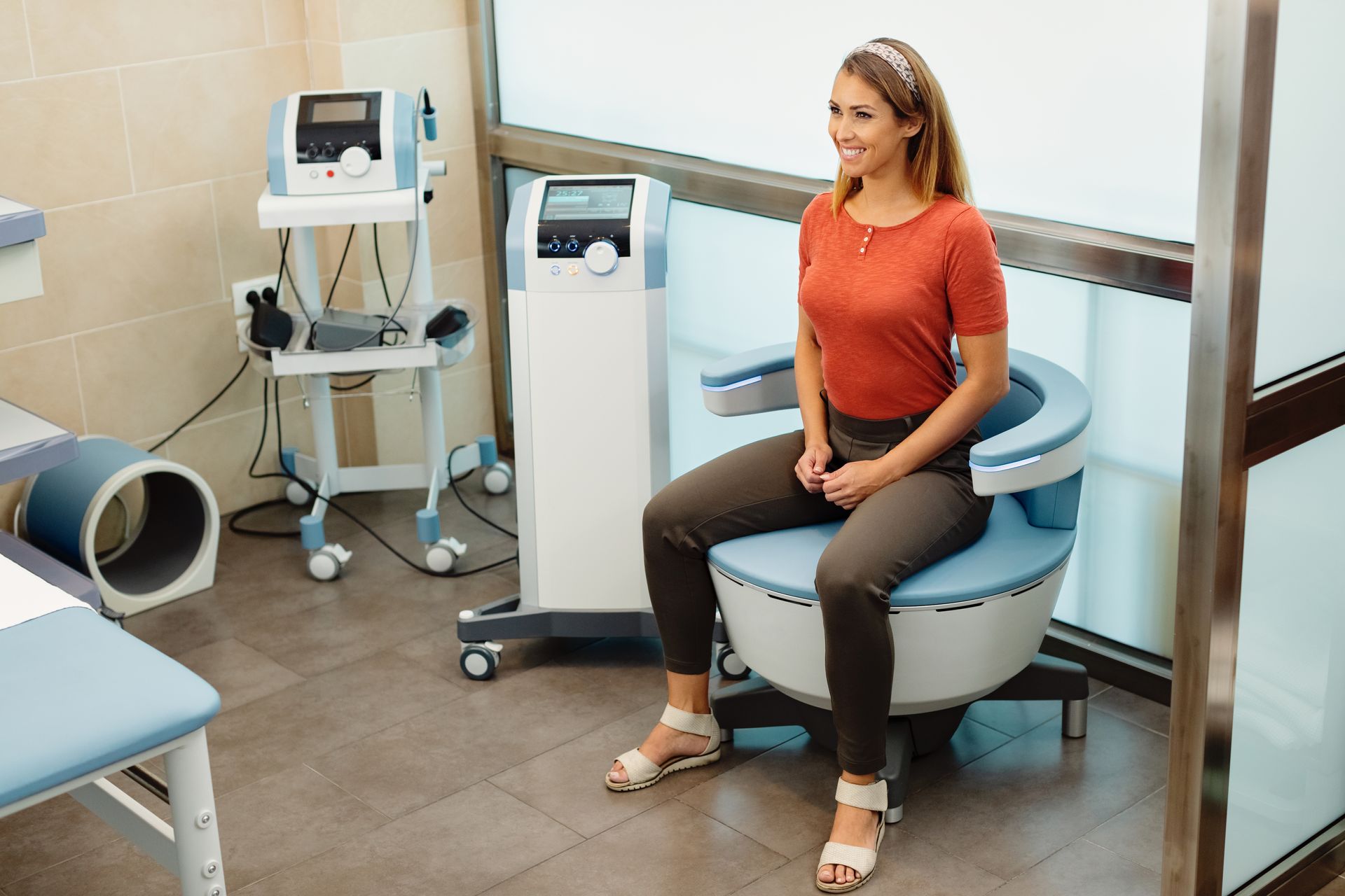 Woman seated on a blue medical chair in a clinic room, smiling beside diagnostic equipment.