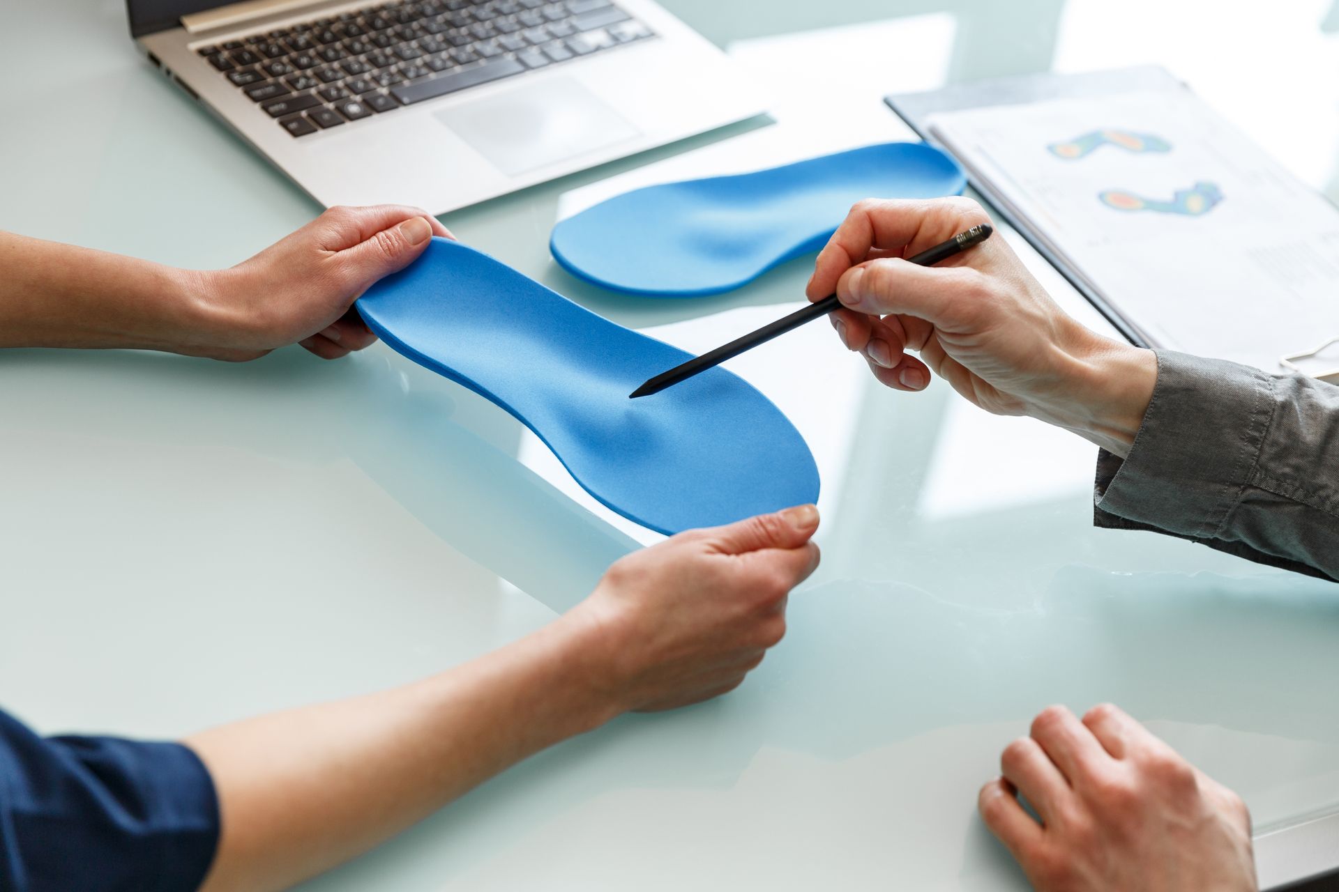 Hands measuring a blue orthotic insole on a desk with a laptop and design tools