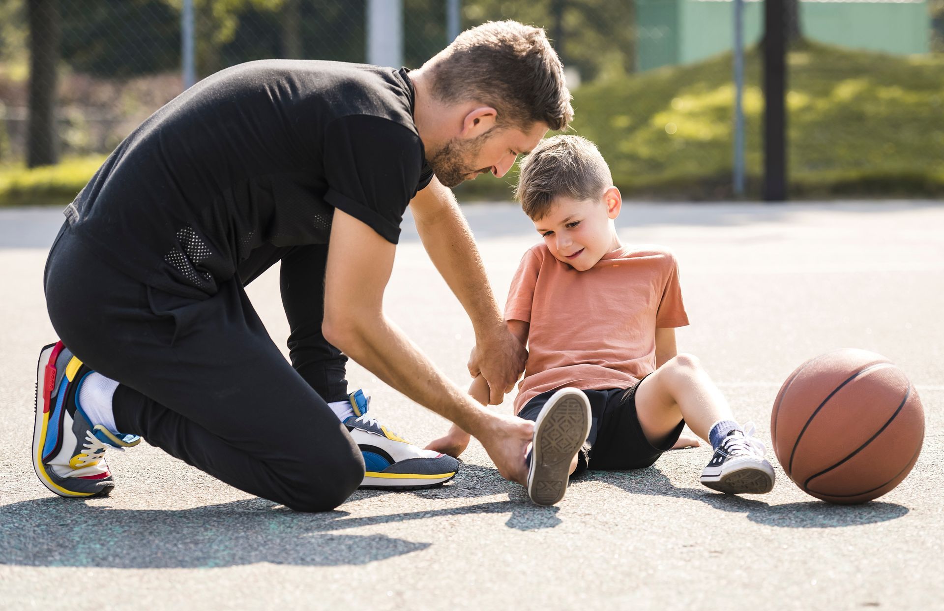 Adult kneeling beside child outdoors, helping them with a basketball on the pavement.