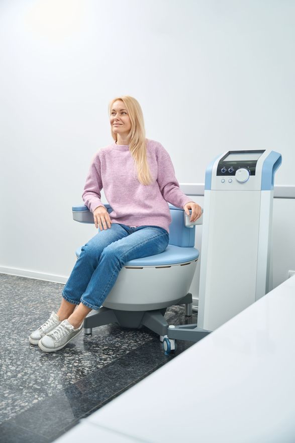 Woman seated on a blue-gray medical chair, wearing a white blouse and black pants, smiling against a white background