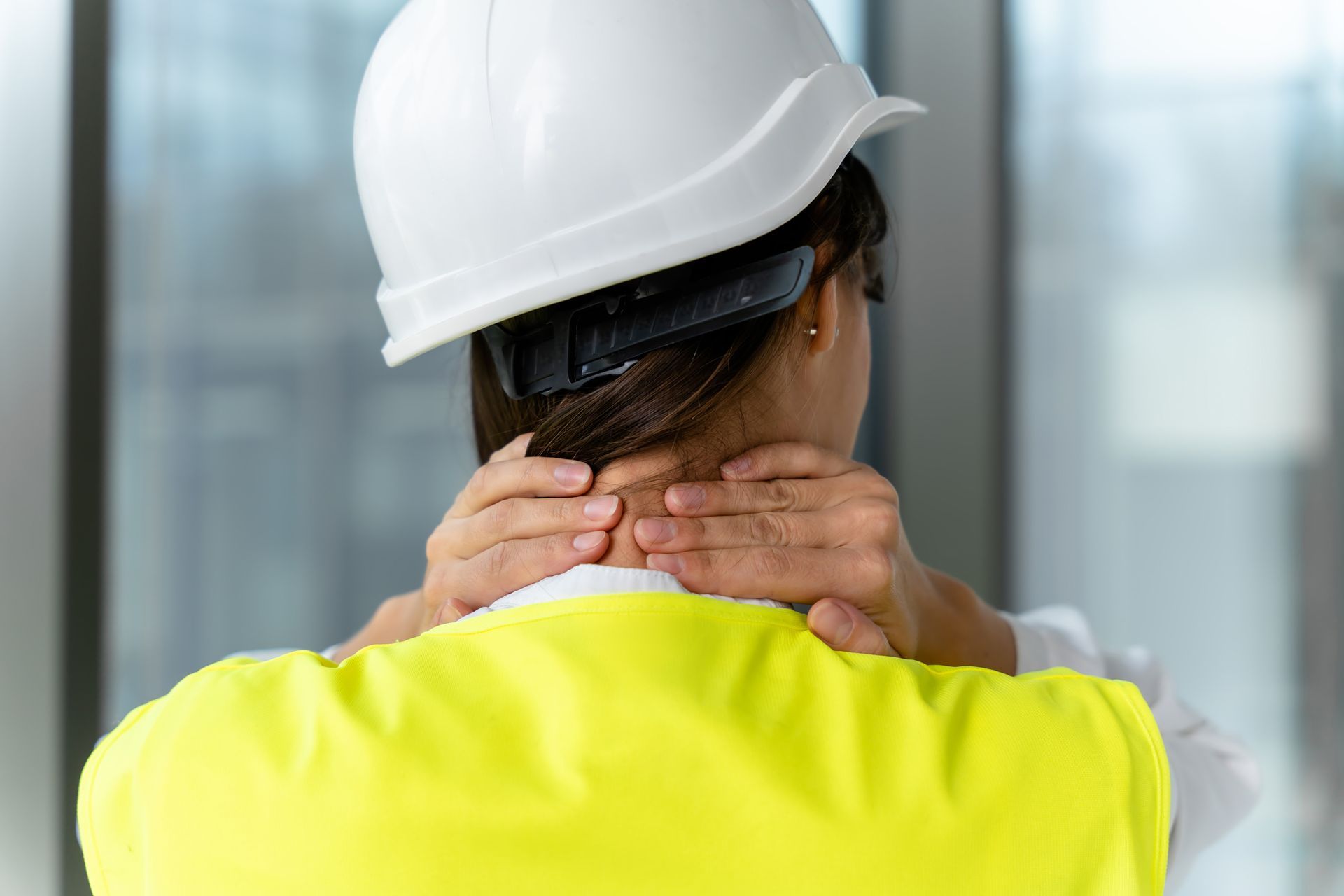 Worker in white hard hat and yellow vest rubbing their neck, seen from behind