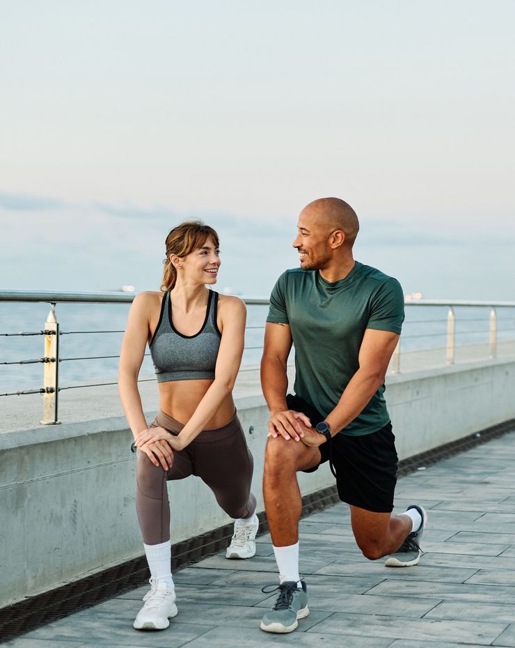 Two people in workout clothes lunging on a waterfront path at sunset