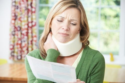 Woman in green sweater wearing a neck brace, reading a document at home with a pained expression