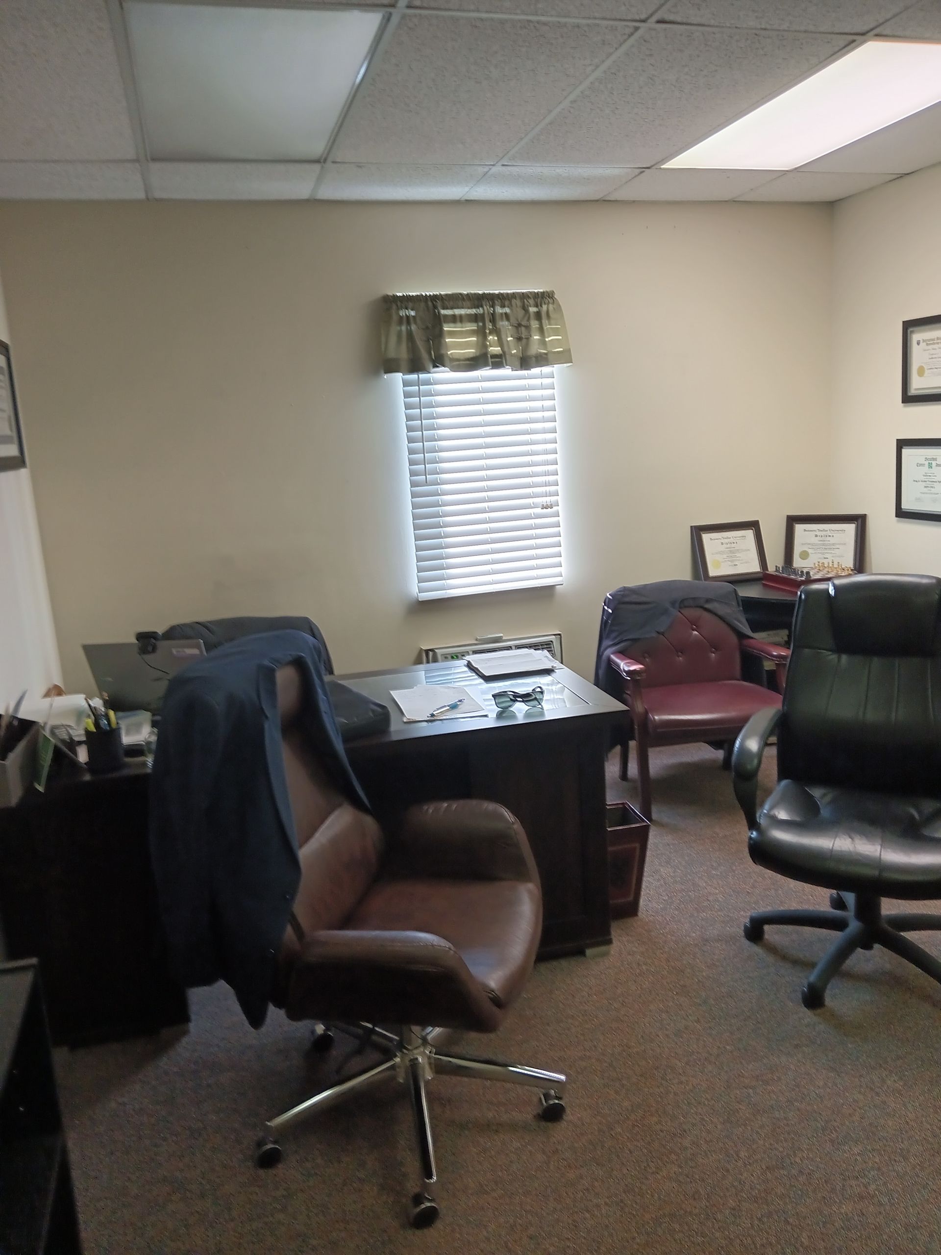 Office with desk, chairs, window with blinds, awards on wall; carpeted floor.