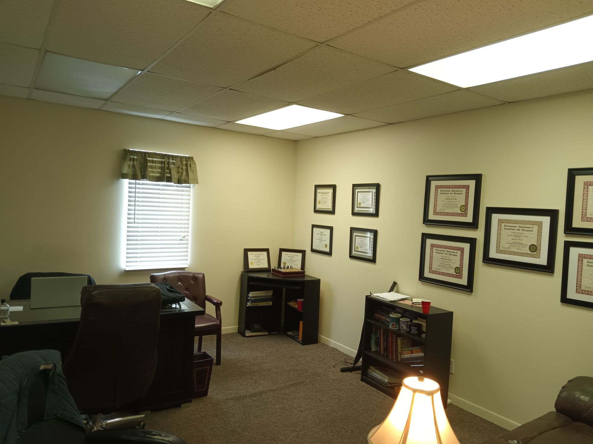 Office with desk, window with blinds, framed documents on wall, and lamp.