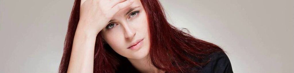 Woman with red hair, hand on forehead, looks troubled. Black top on, neutral background.