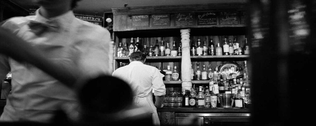 Bartenders at work in a dimly lit bar, bottles on shelves behind them.