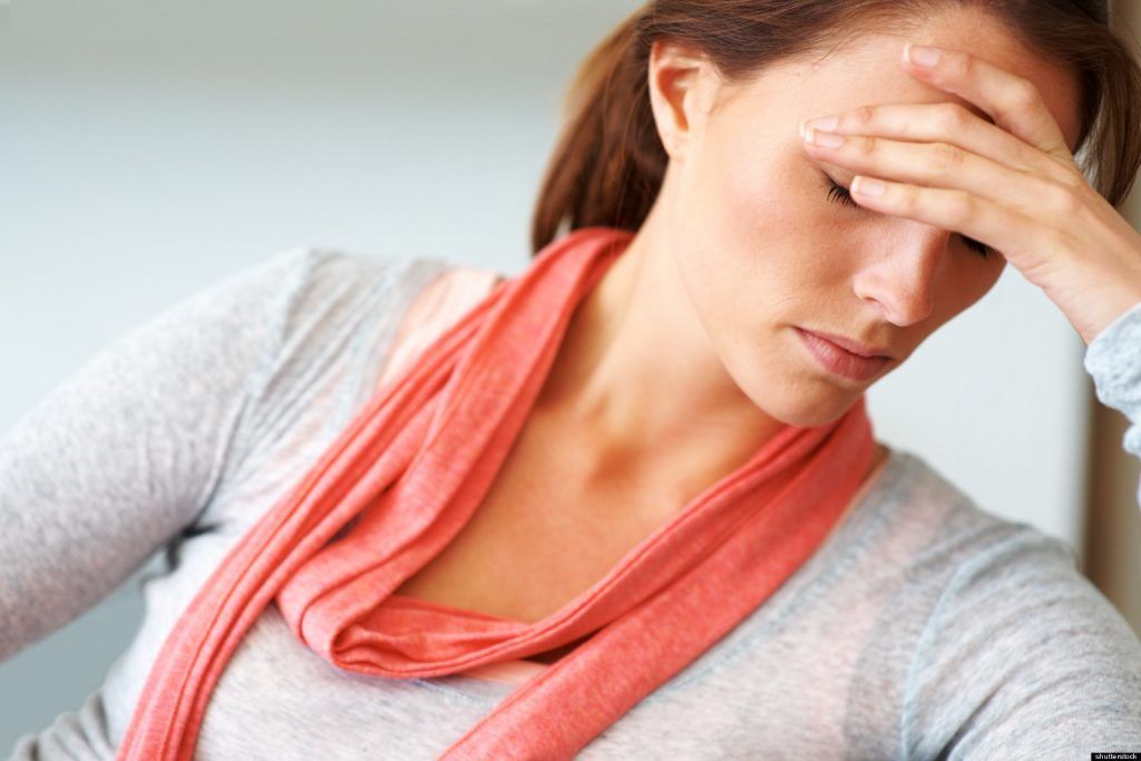 Woman with hand on forehead, looking down, appearing stressed or in pain. Wearing grey top and coral scarf.