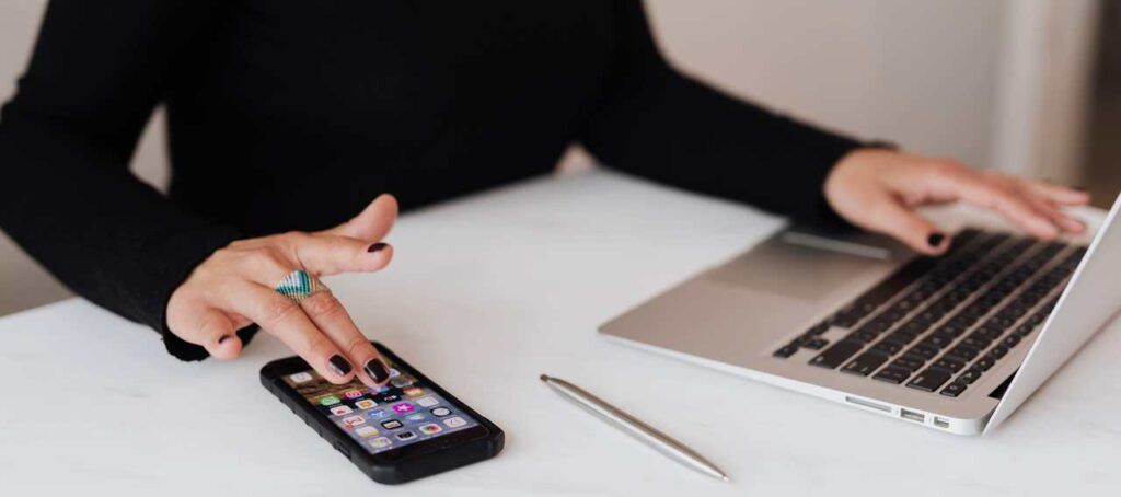 A person with black nail polish uses a phone and laptop on a white desk. There is also a silver pen.