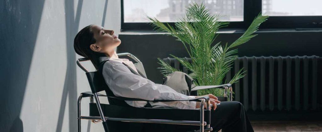 Person reclines in chair, head tilted toward sunlight. Indoor setting with a plant.
