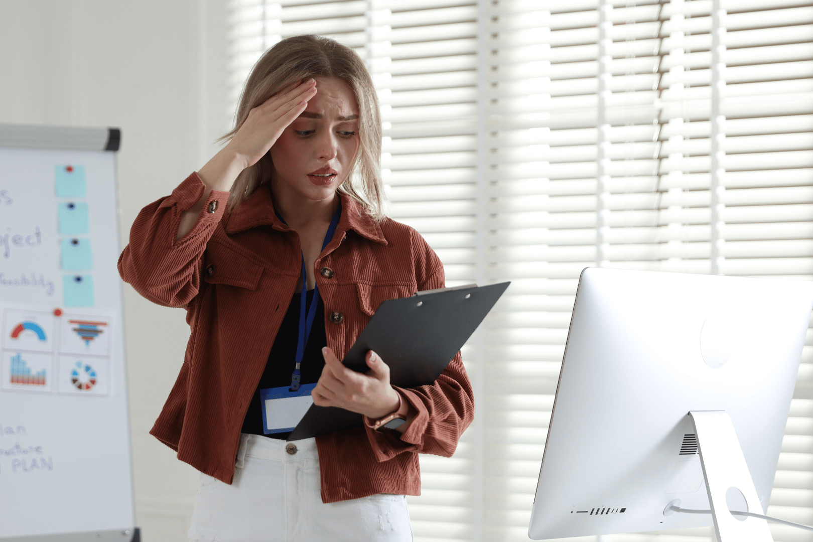 Woman in office with hand on forehead looking stressed, holding clipboard near computer.