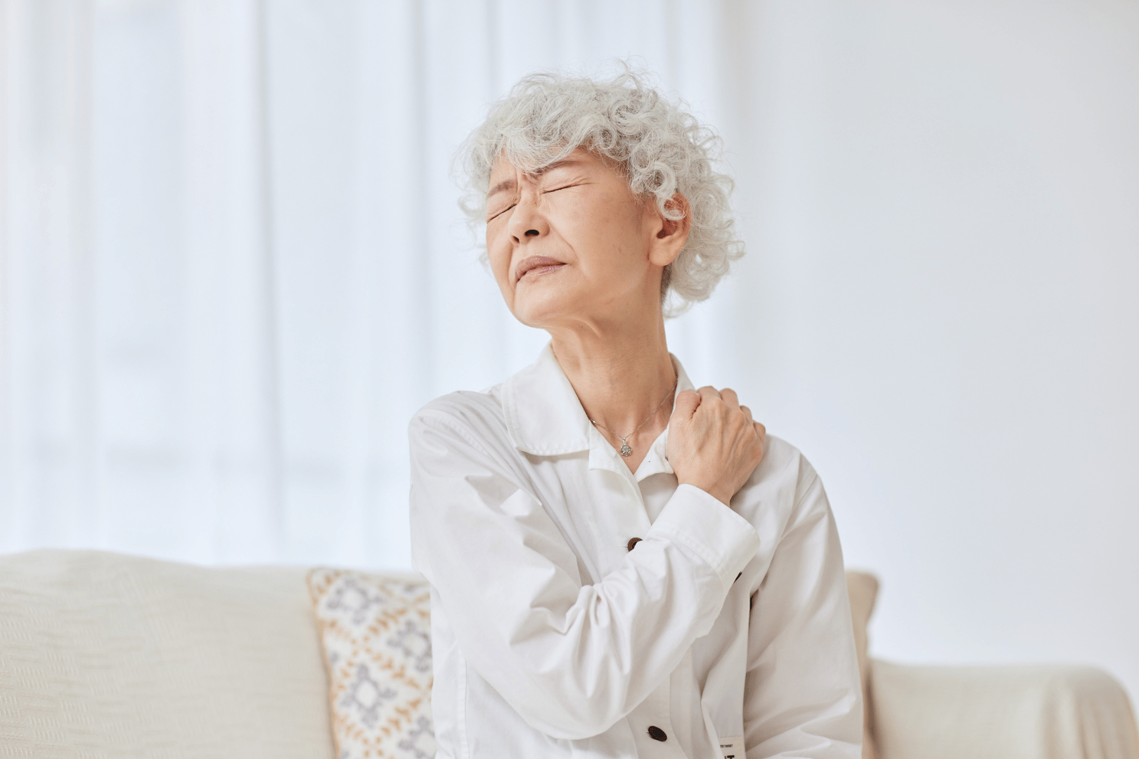 Woman holding her shoulder, looking pained. She is indoors, possibly on a couch.