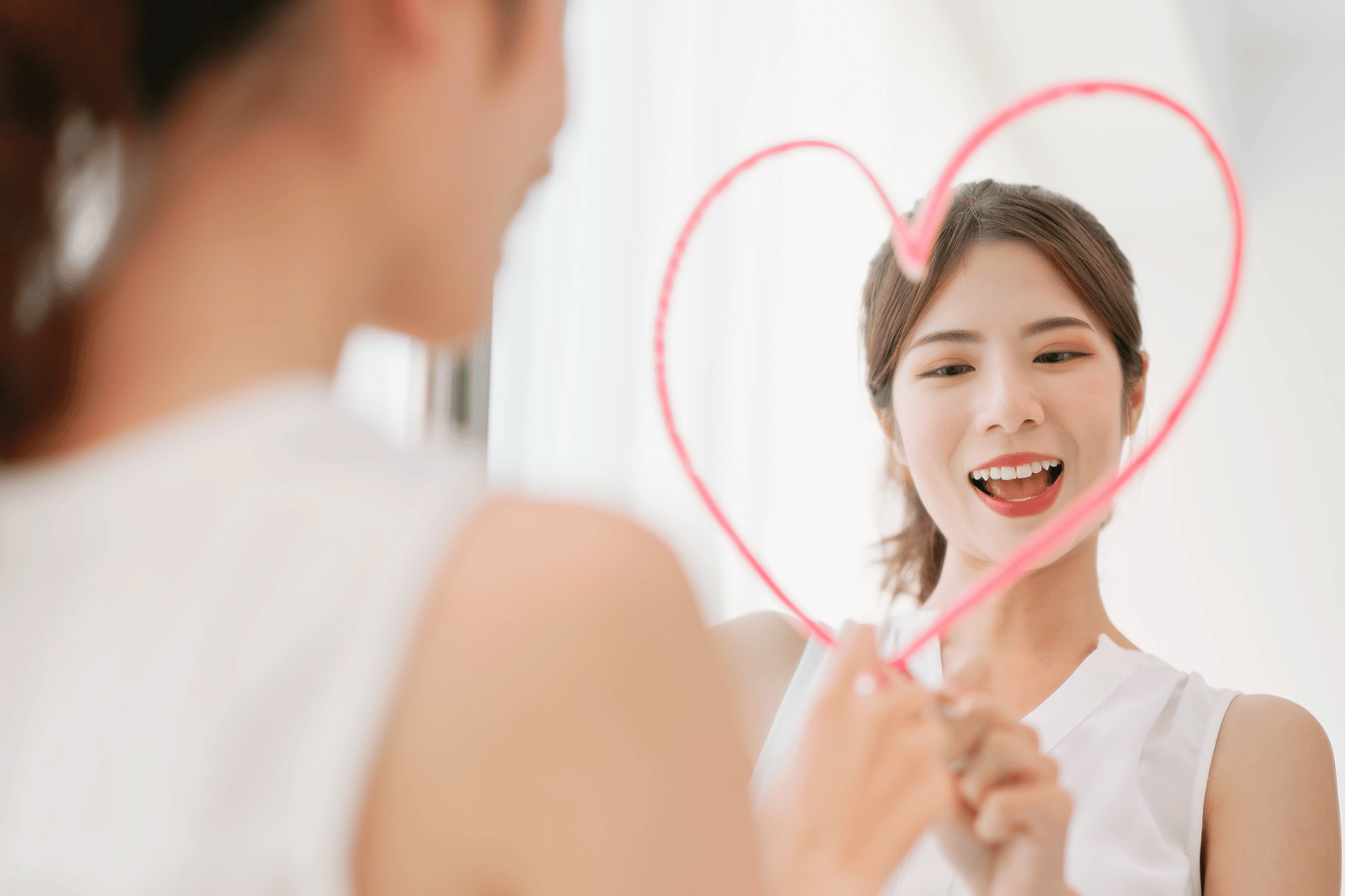 Woman holding a pink heart-shaped frame smiles at her reflection in a mirror.