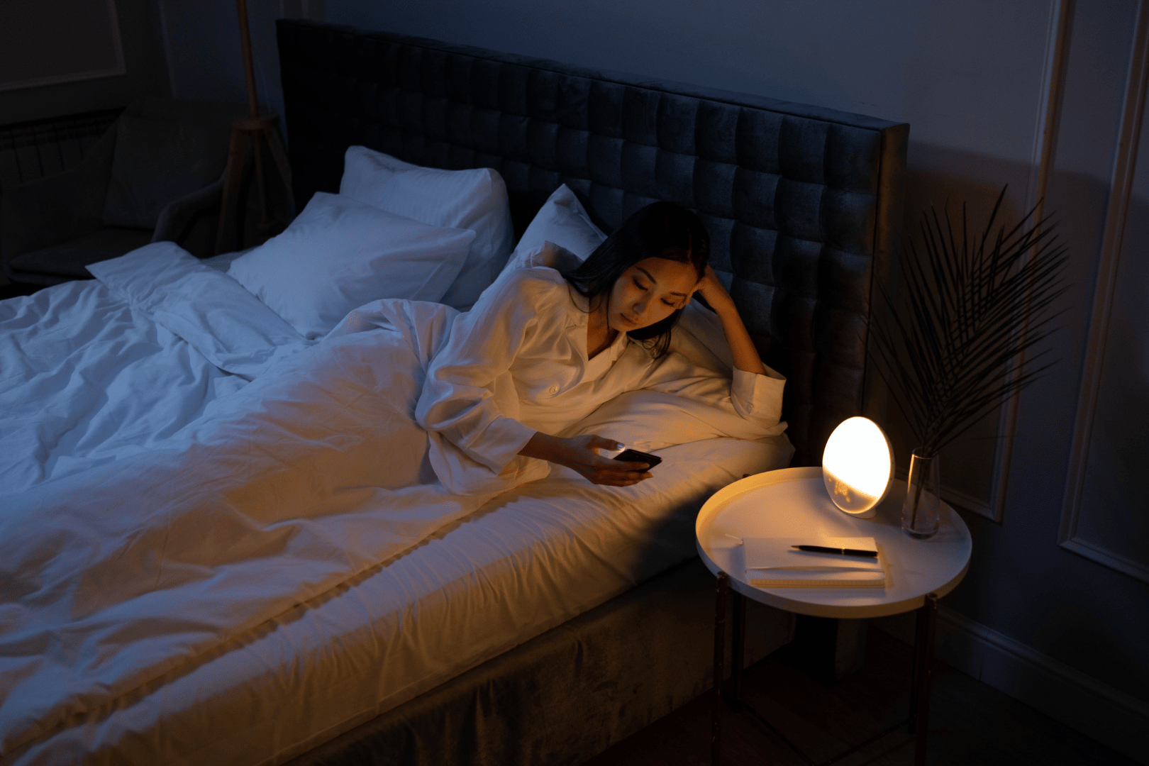 Woman in bed, looking at a phone, lit by a bedside lamp.
