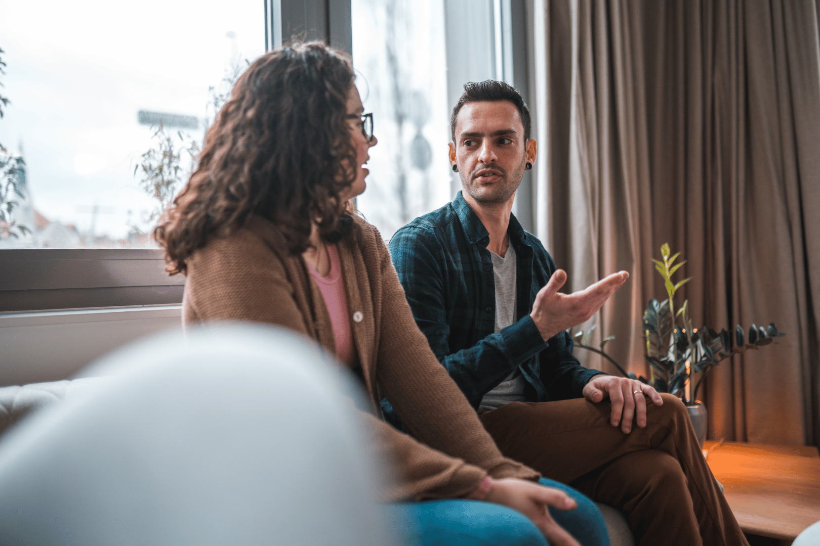 Couple sitting and talking, near a window. Man gestures with his right hand. Woman wears glasses and smiles.