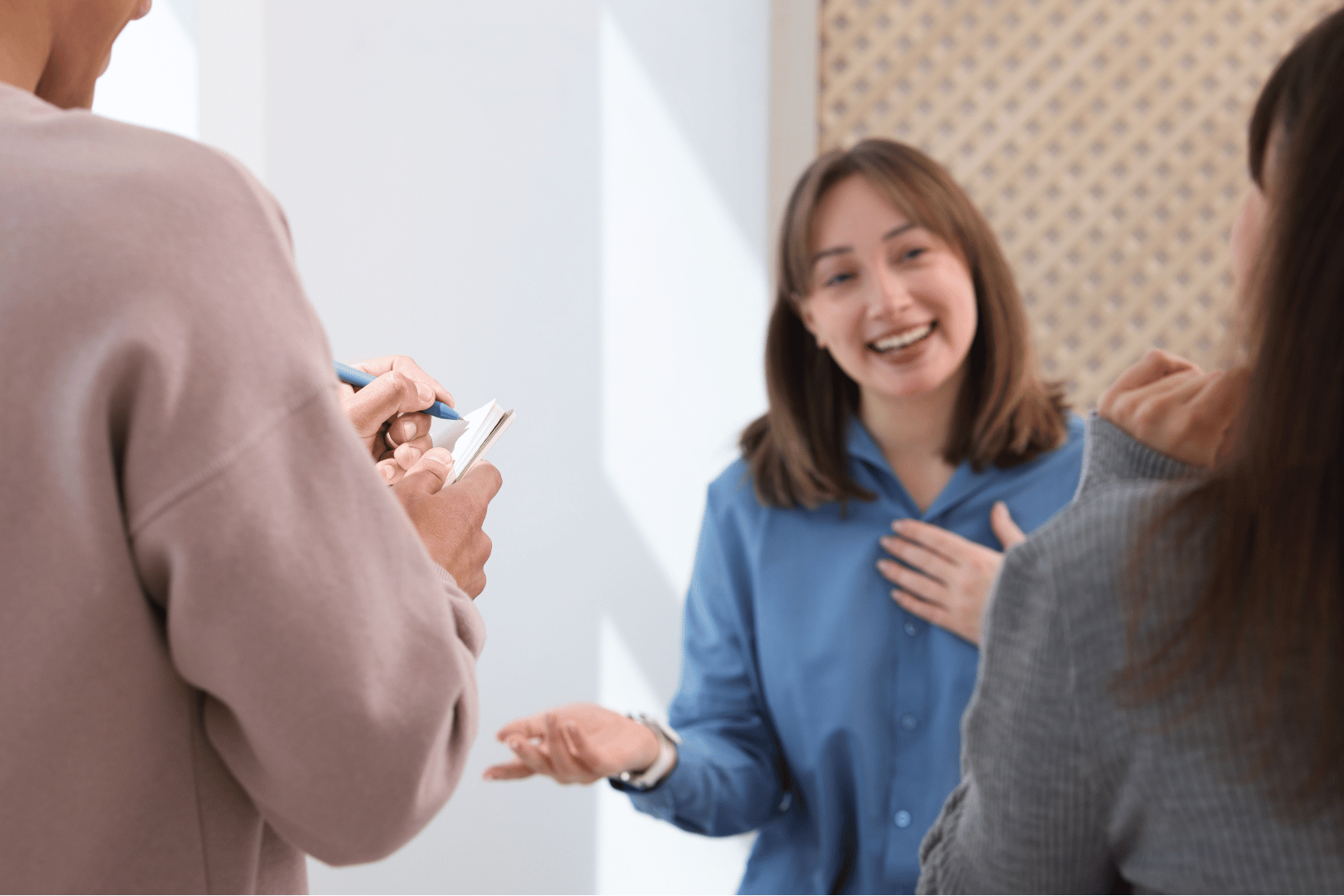 Three people in a bright room. Woman in blue shirt smiles, gesturing. Someone takes notes.