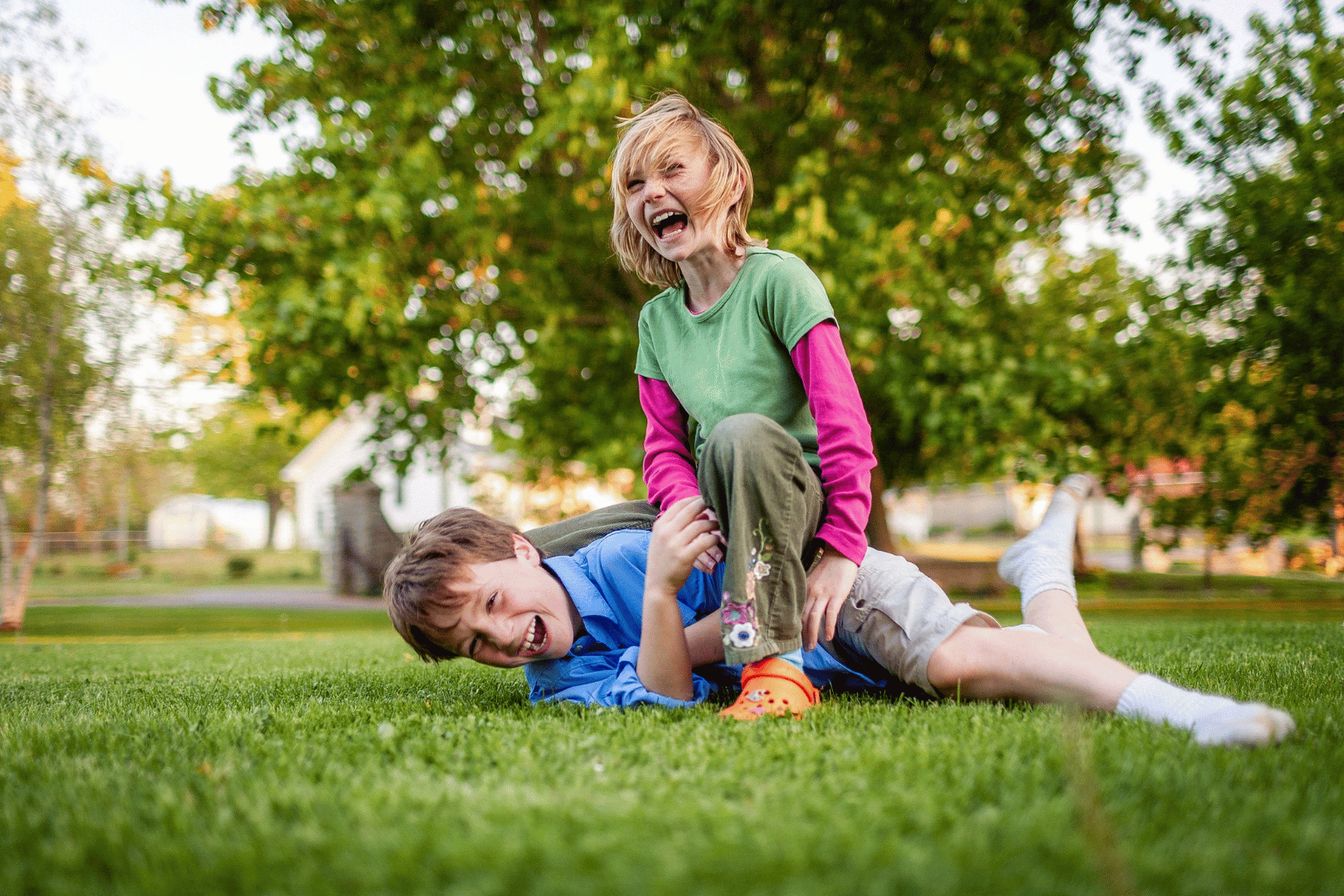 Two children playing on green grass, one laughing on top of the other, near a tree and a building.