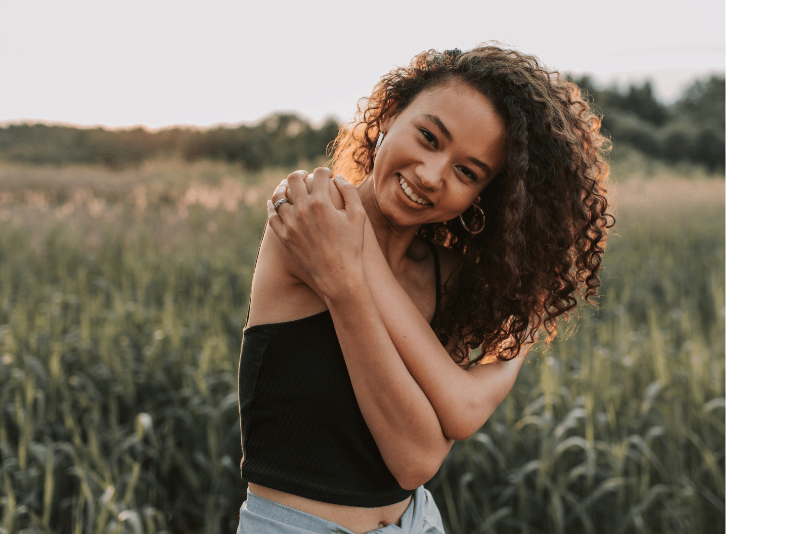 Woman with curly hair hugging herself, smiling in a field of tall grass during sunset.