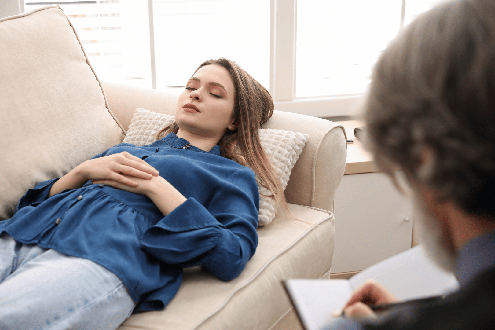 Woman lying on a couch with eyes closed; a person with notepad is seated nearby.