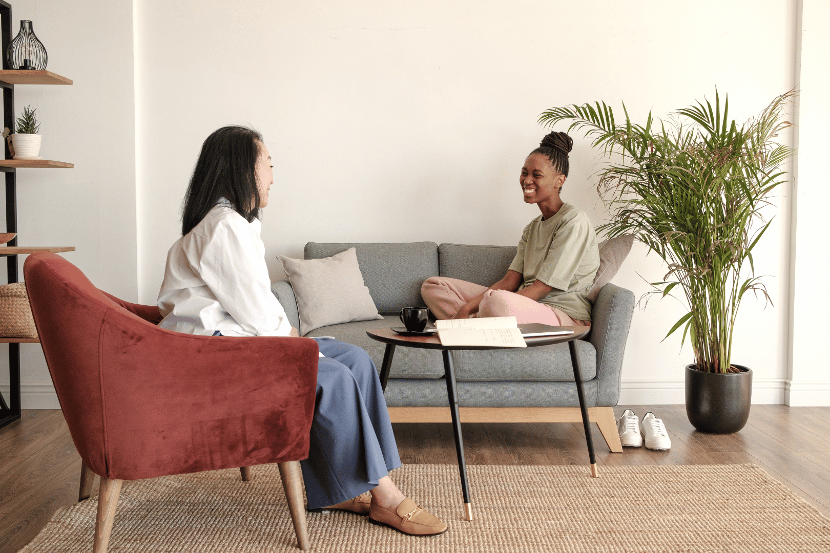 Two people in a therapy session: one sits on a couch, the other in a chair, a plant beside them.