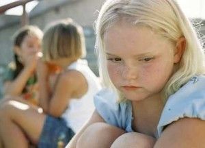 Blond girl looks sad while two others whisper nearby. Outdoor setting, daylight.