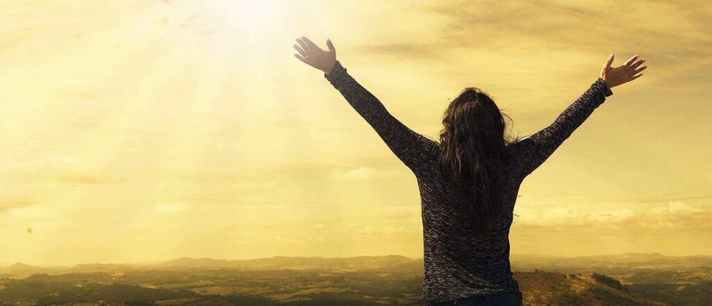 Woman with arms raised, silhouetted against a bright, golden sky over a landscape.
