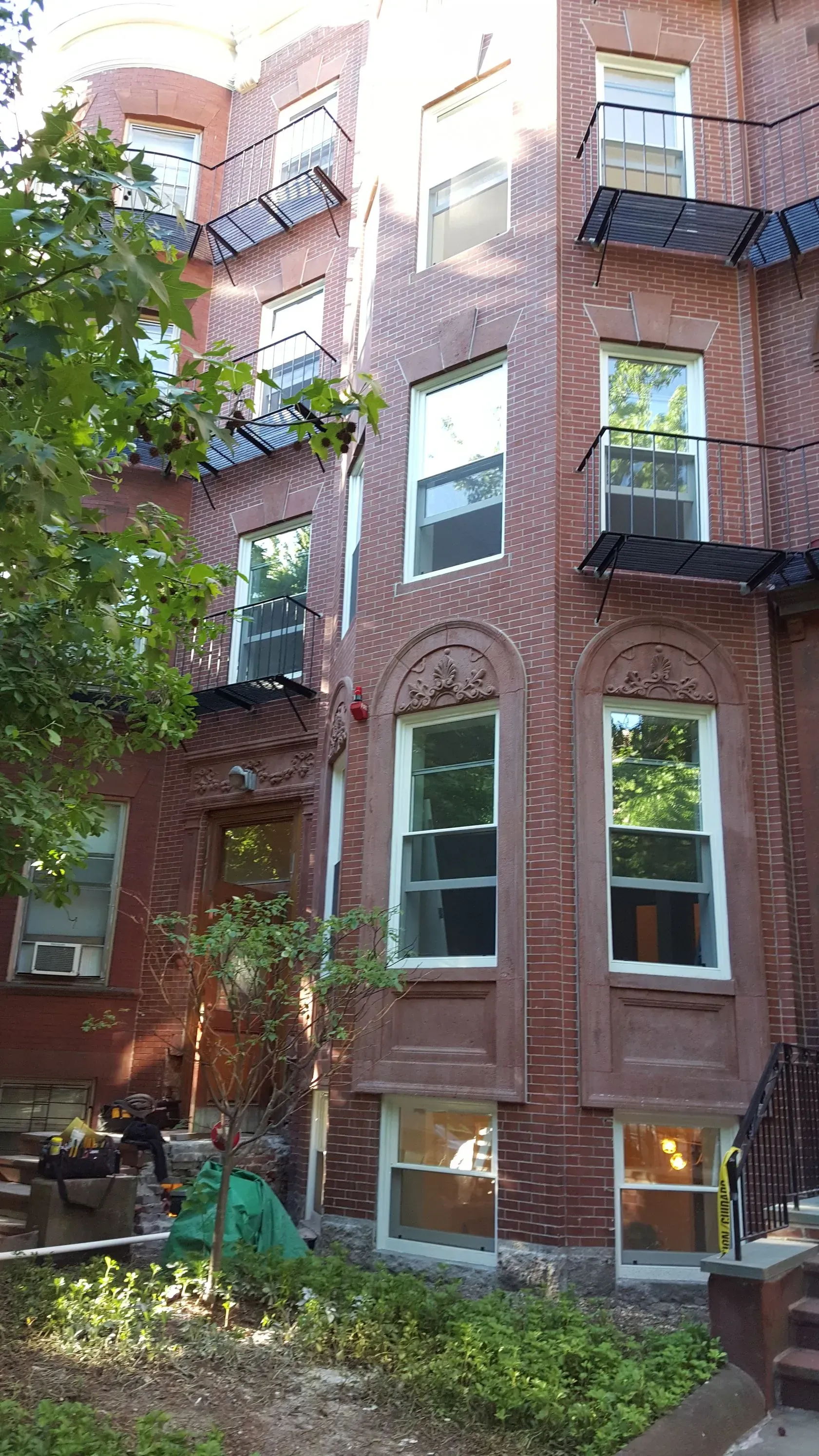 Red brick building with multiple windows and small balconies. Green foliage in foreground.