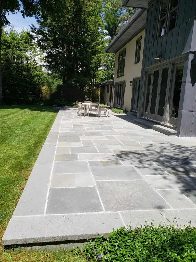 Stone patio beside a house with a table and chairs in the distance; green lawn and foliage.