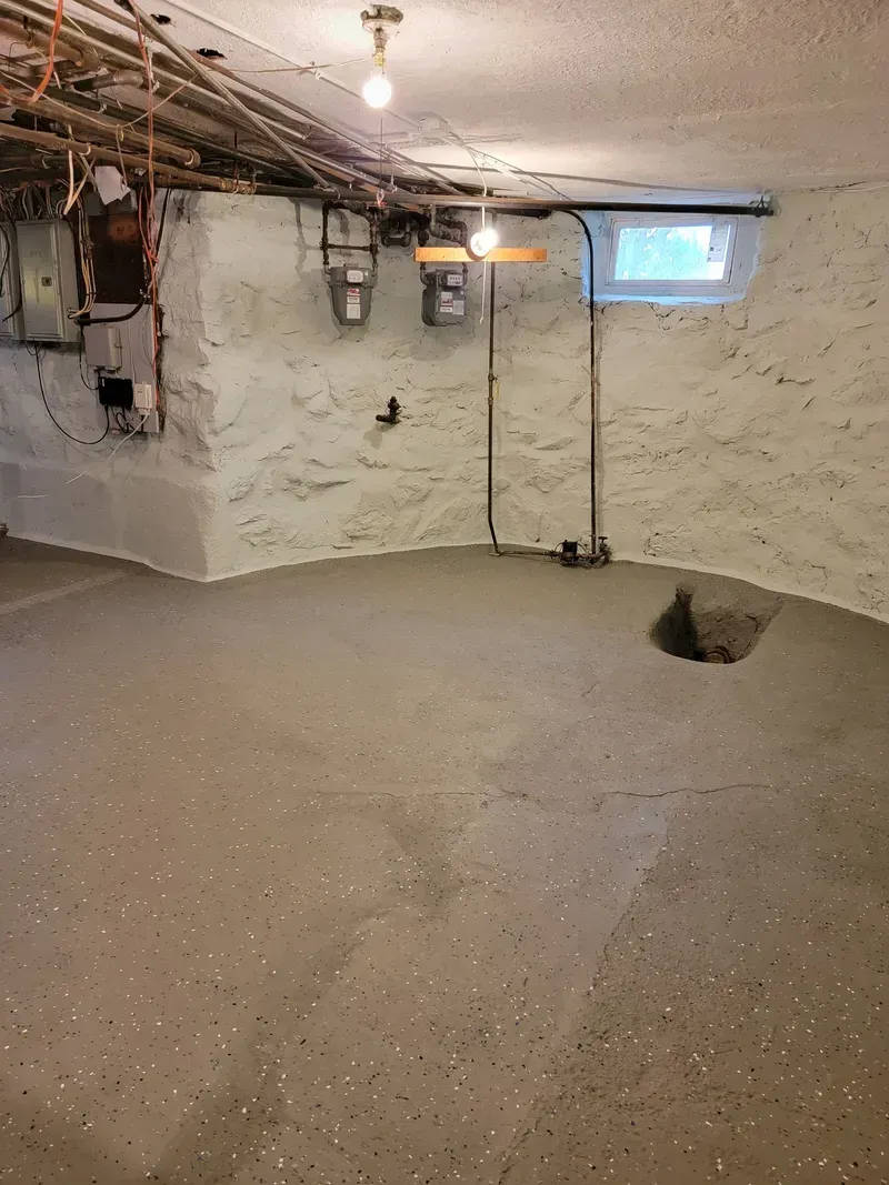 Basement with rough concrete floor, white-painted stone walls, pipes, and small window.