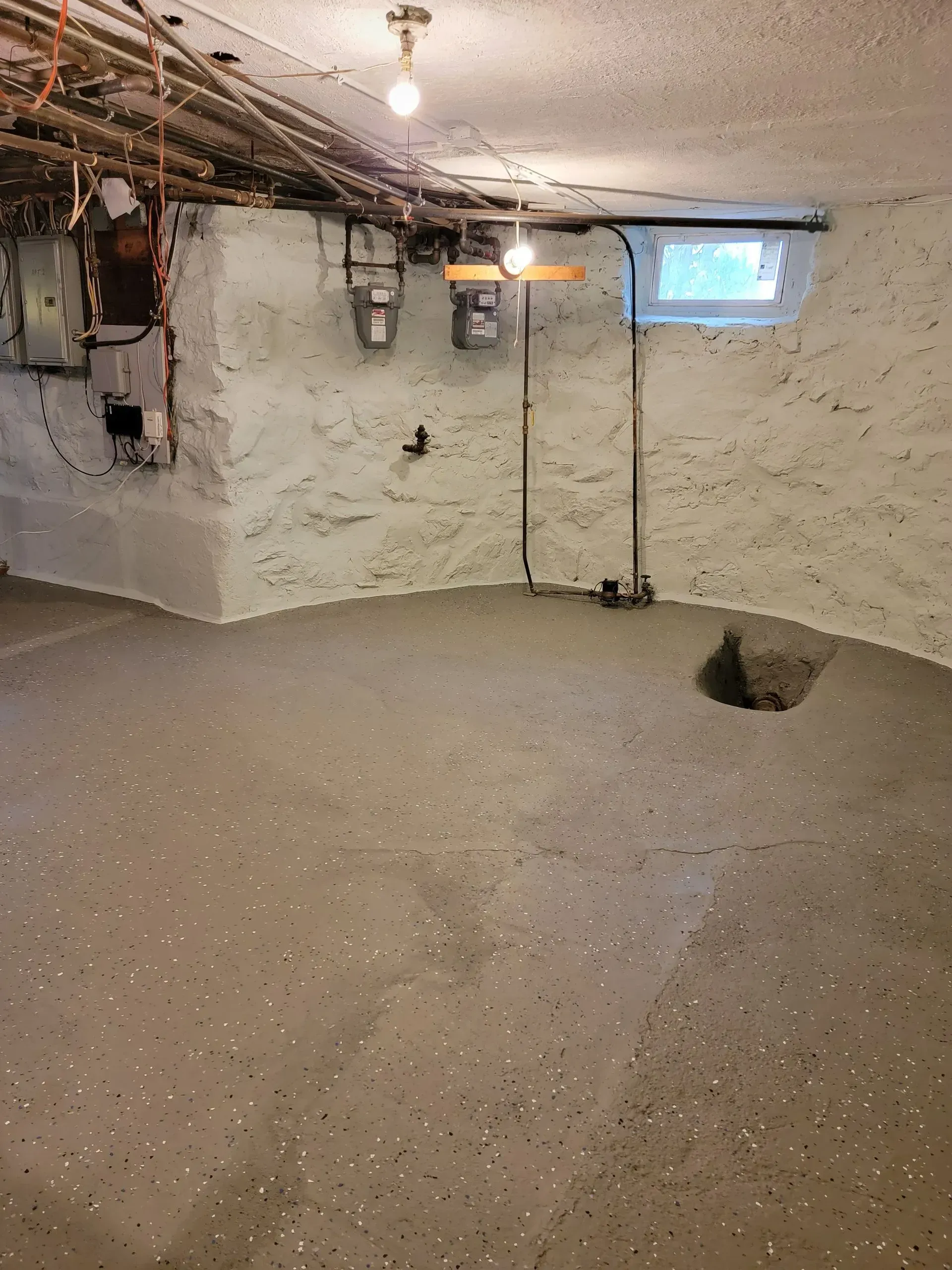 Basement with concrete floor, white textured walls, and exposed pipes and utilities.