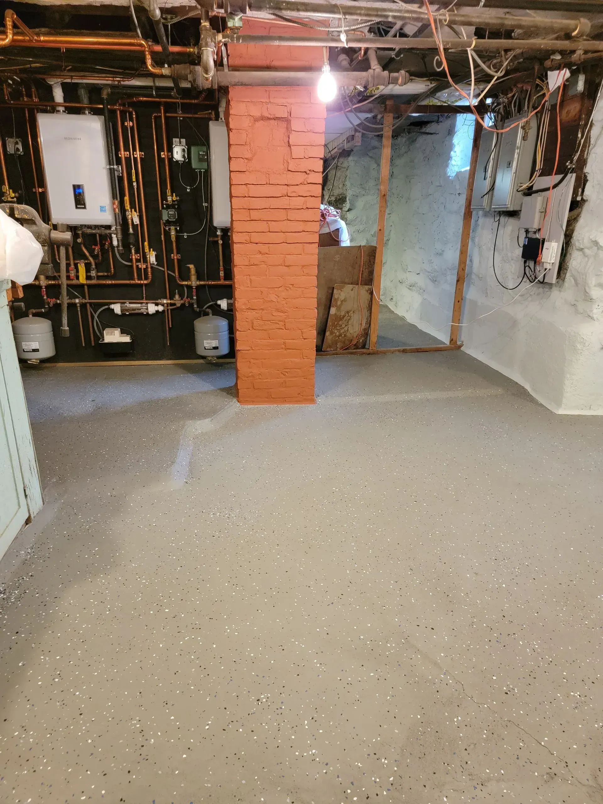 Basement with painted gray floor, brick support beam, and exposed pipes on the ceiling.